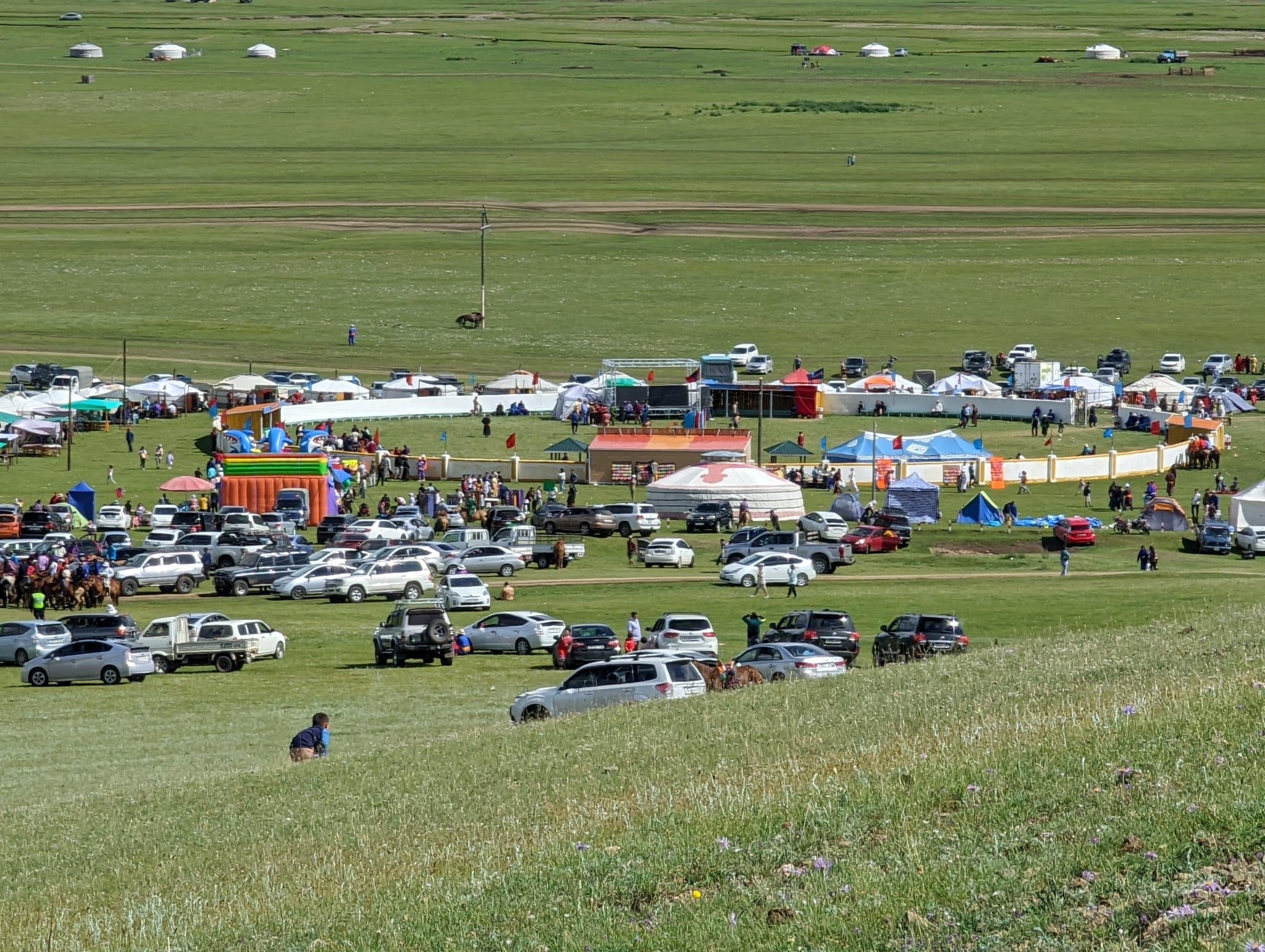 A village Naadam ground — gers, market tents, a bouncy castle, parked cars, and the roped arena on the open steppe
